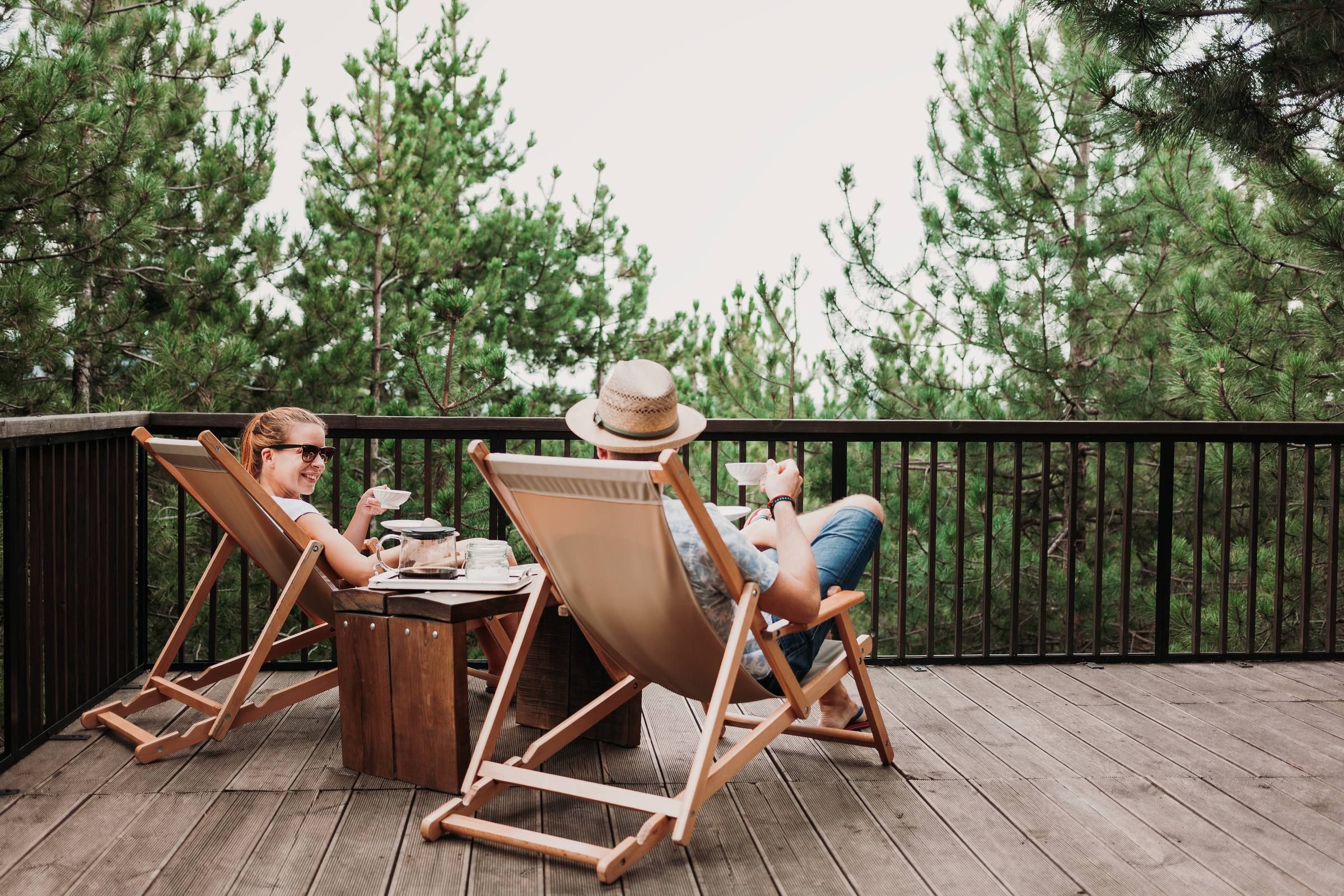 Two people relaxing on deck chairs on a wooden patio, surrounded by trees, enjoying drinks and snacks.